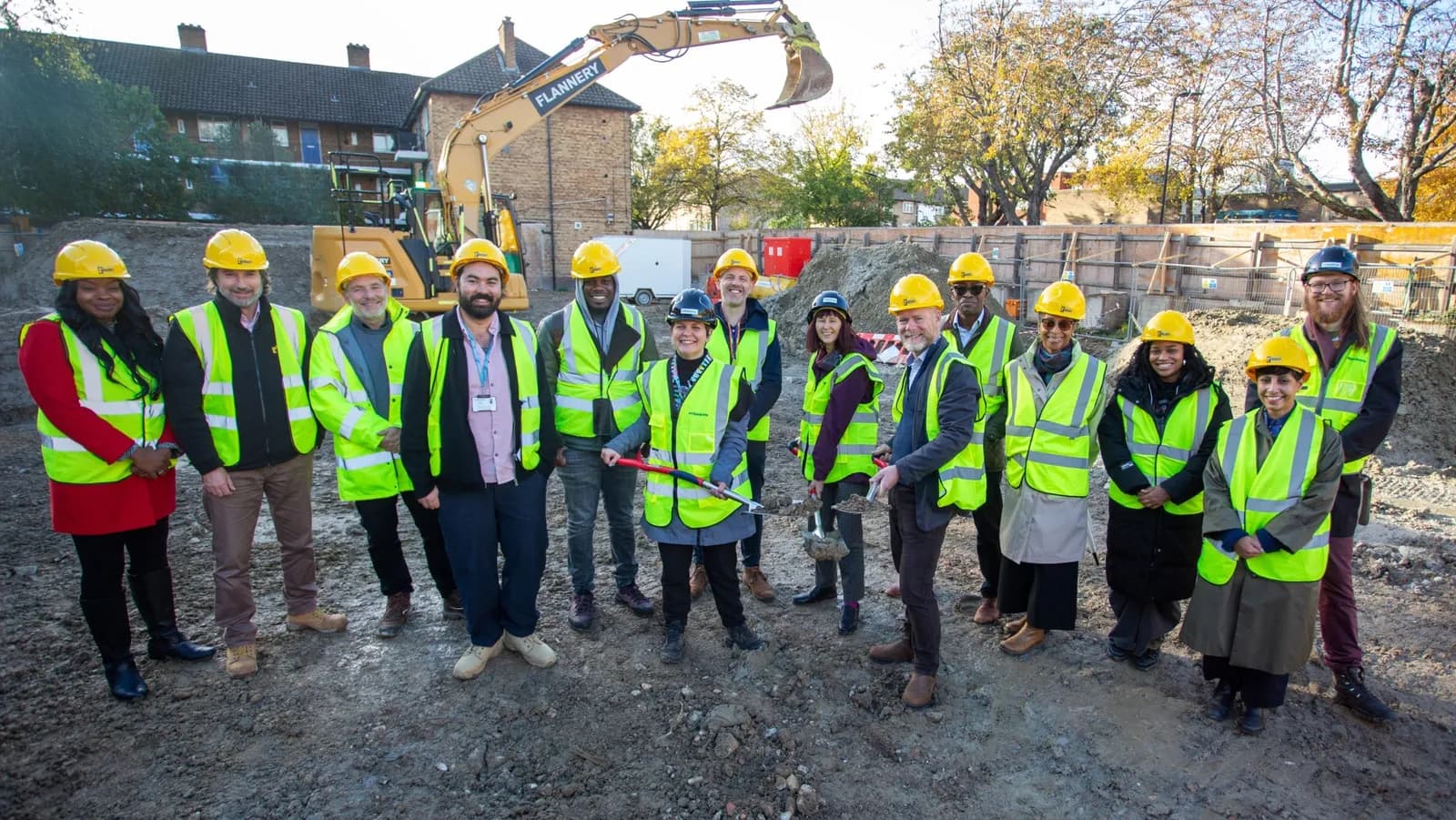 A diverse group of 14 people wearing yellow hard hats and high-visibility vests smile while standing on a construction site with an excavator in the background.